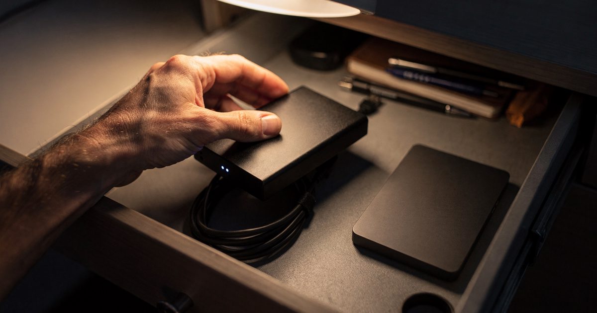 Tight overhead close-up of an open desk drawer under warm lamplight at night, a hand lifting a scuffed external hard drive off its coiled USB cable while a.