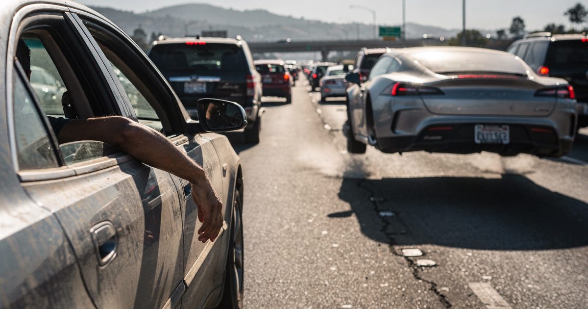 Low-angle medium shot in harsh midday sun on a jammed multi-lane California freeway, a commuter's arm resting out the open window of a dusty sedan in the.