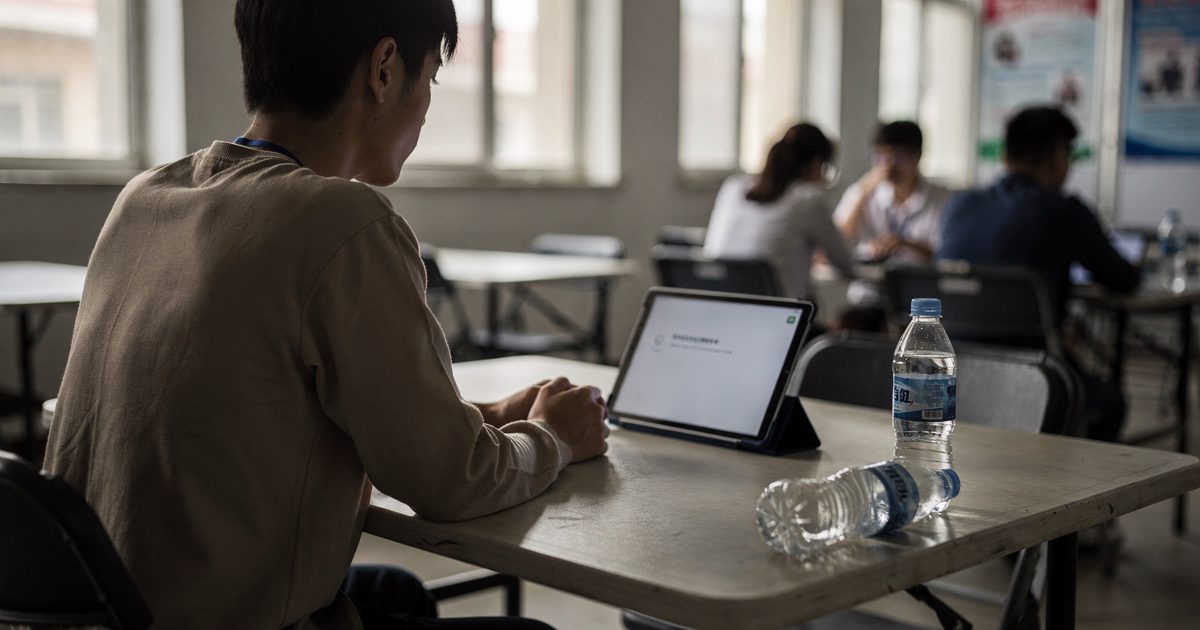 Mid-morning sun through high windows of a campus exhibition hall as a graduate cropped at the neck sits across a folding table from a tablet, an empty water.