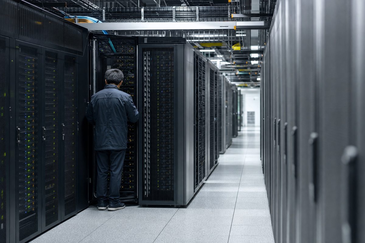 Wide photojournalistic image inside a modern server room, one technician seen from behind checking a rack of blinking equipment, cool industrial lighting, no.