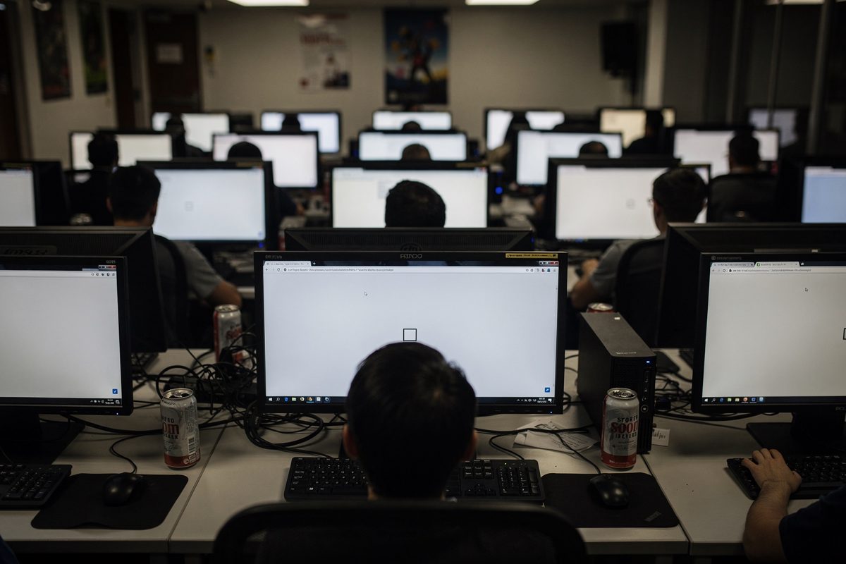 Wide shot across a university computer lab under flat fluorescent light at dusk, rows of students seen from behind at identical monitors, every screen frozen.