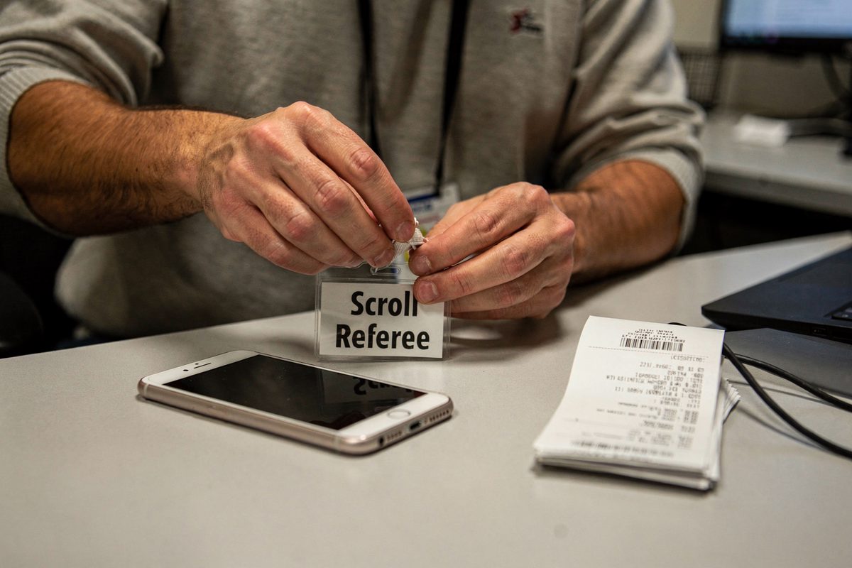Harsh afternoon fluorescent light on an office desk where a pair of hands clips a badge reading "Scroll Referee" onto a cloth lanyard, a face-down test phone.