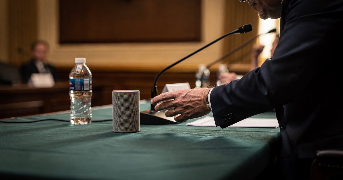 Close detail in warm late-afternoon light slanting across a Senate hearing room witness table, a small grey smart speaker standing on the green baize beside a.