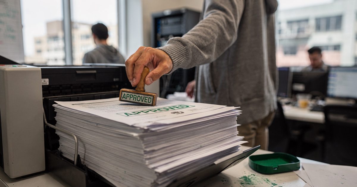 Medium shot in bright mid-morning window light at a printer alcove inside a San Francisco tech office, a staffer seen from behind pressing a rubber stamp.
