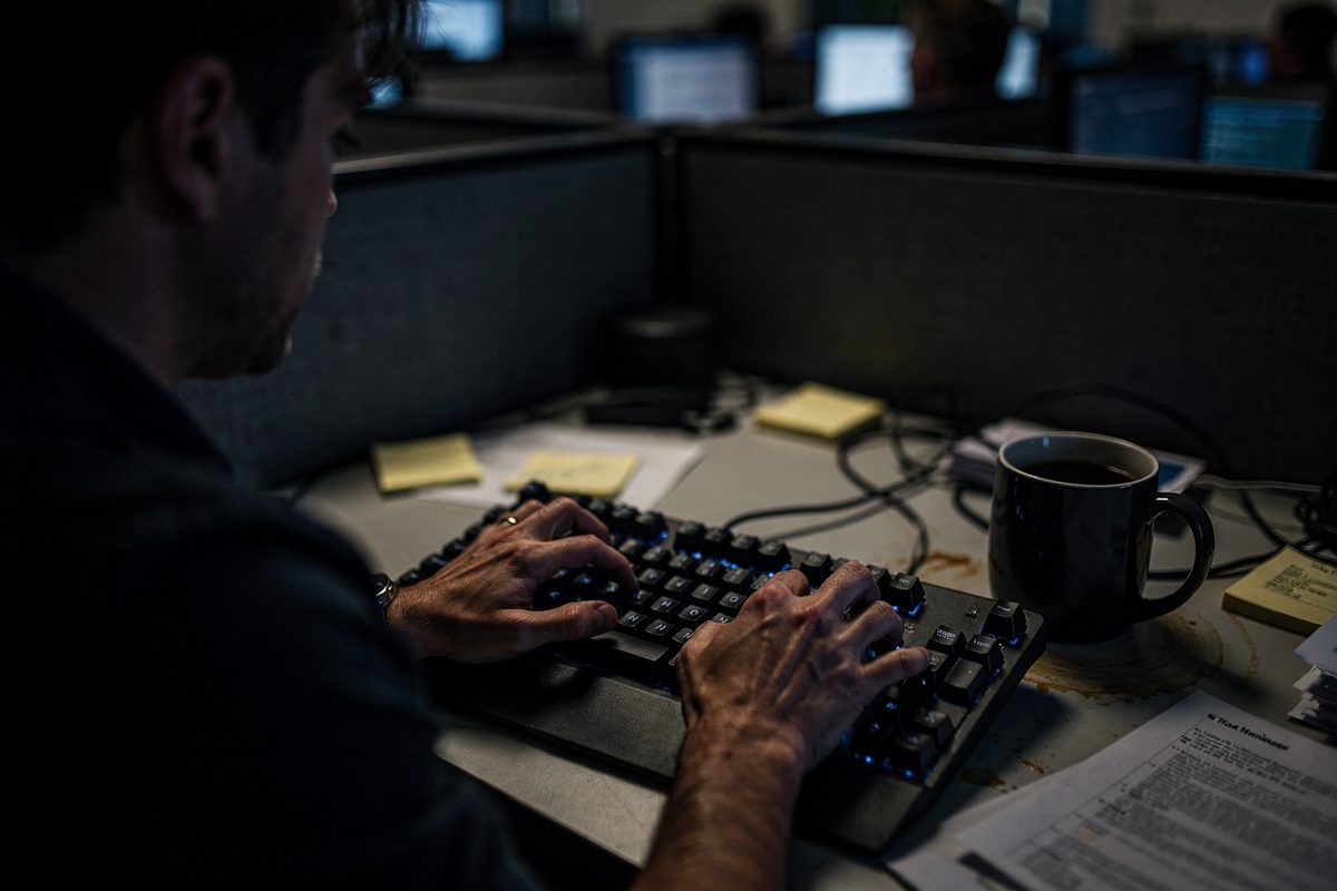 Close-up over the shoulder of a frantic typist in a dimly lit cubicle, showing hands blurred in motion above a mechanical keyboard beside a half-empty mug of.