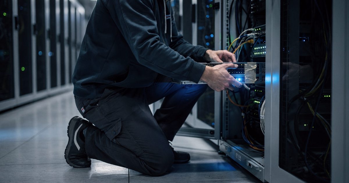 Cool blue LED lighting inside a dense Frankfurt data center, a network engineer seen from the waist down kneeling to unplug a glowing server blade labeled.