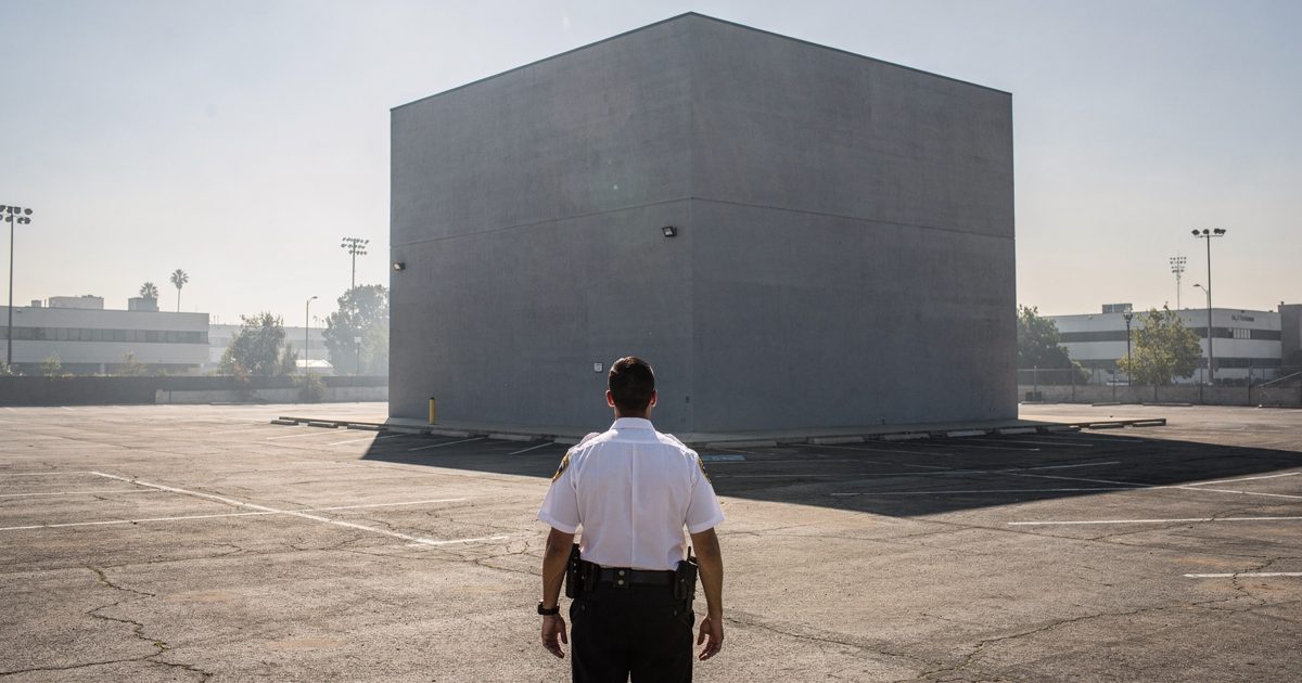 Wide shot in harsh afternoon sunlight of a vast empty asphalt parking lot in Burbank, a massive, seamless gray steel cube sitting alone in the center while a.