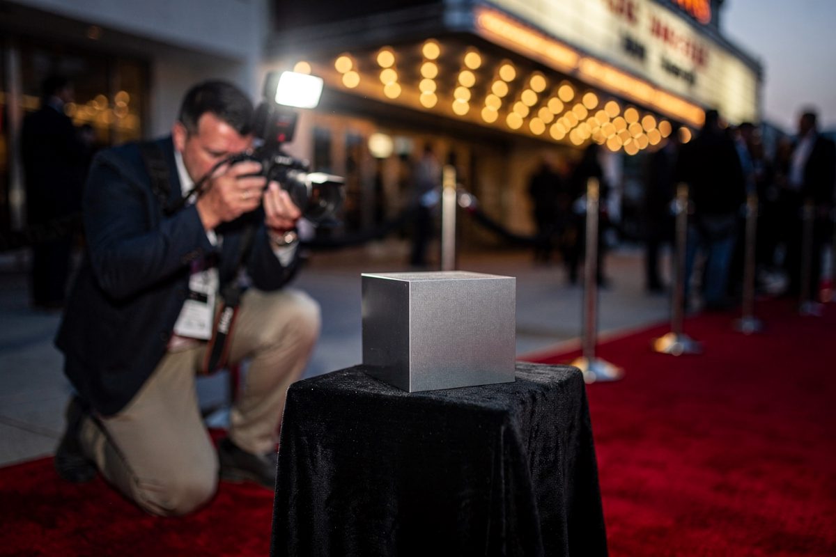 Medium shot at dusk on a red carpet outside a Los Angeles theater, a photographer in a dark jacket kneeling to aim a camera equipped with a large flash at a.