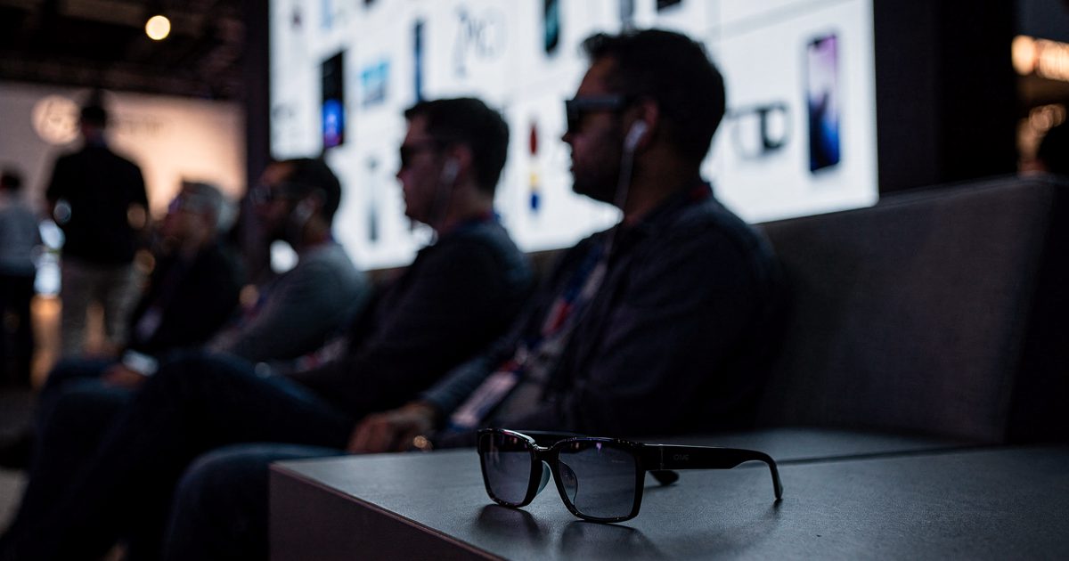 Mid-shot at dusk in a tech conference demo lounge lit by a glowing product wall, a row of attendees seen in silhouette from the side each tilting their head.