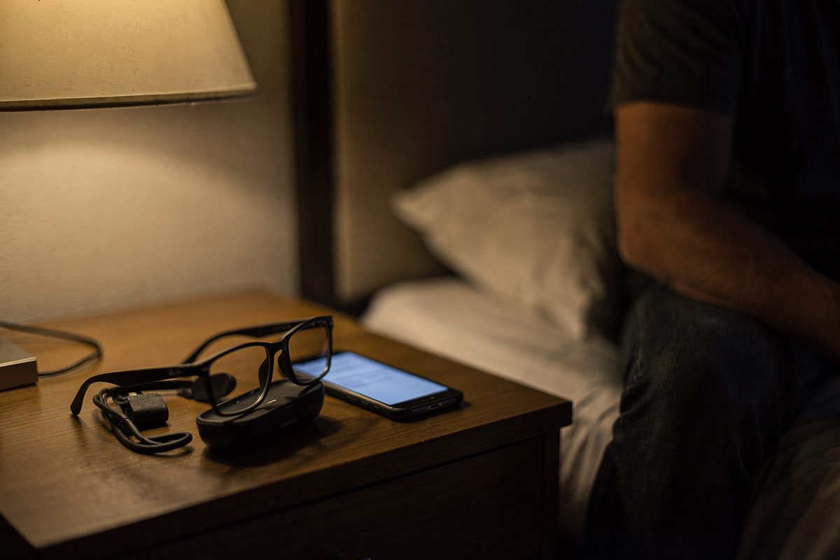 Close detail at night on a hotel nightstand under a single bedside lamp, a pair of dark-framed smart glasses set down beside a phone whose screen glows with an.
