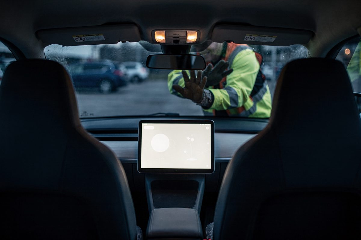 Pre-dawn blue light inside a small two-seat car with no steering wheel, the dashboard screen glowing over two empty seats, and a plant inspector's face blurred.