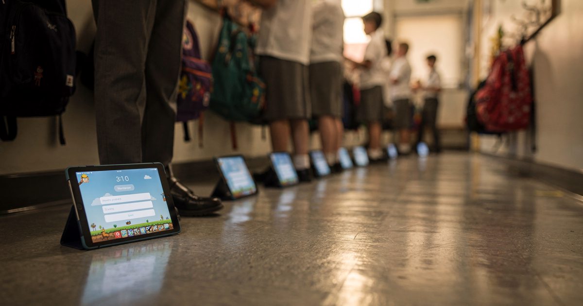 Low shot from floor level down a primary school corridor in warm late-afternoon light, a row of children in grey uniform trousers seen only from the knees.