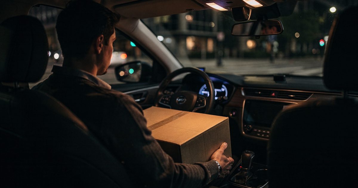 Cabin light at early night inside a robotaxi as a passenger seen from behind in the rear seat steadies a cardboard moving box on their knees, the empty.