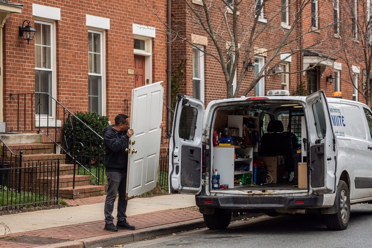 Wide shot in flat noon light on a Washington street of brick rowhouses, a locksmith's van parked with its rear doors thrown open while a man on a stoop, his.