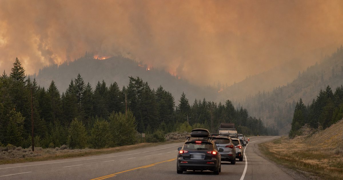 Documentary news photograph of a British Columbia highway under a smoke-filled orange-grey sky, with a line of civilian vehicles moving away from a distant.