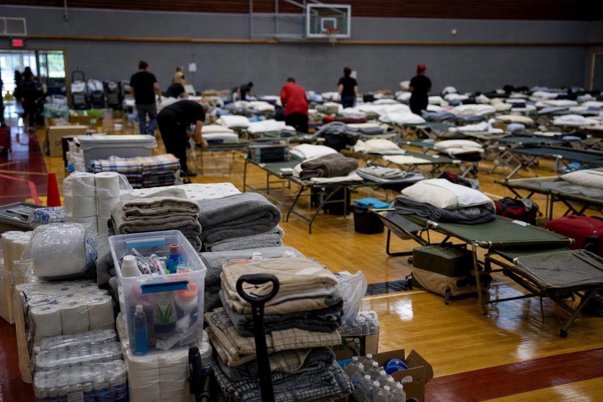 News photograph of an evacuation reception centre in a public gymnasium, rows of cots and stacked supplies prepared by anonymous volunteers, respectful.