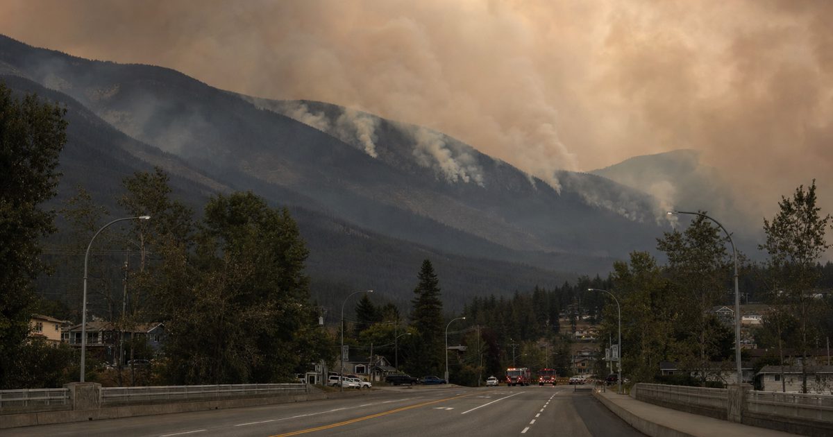 Wide documentary news photograph of a British Columbia mountain community beneath a smoke-filled orange-grey sky, with an empty road, evacuation signage and.