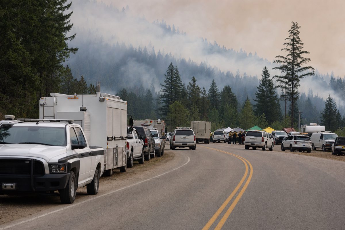 Documentary photograph of unmarked federal and provincial emergency vehicles gathered at a roadside staging area near a smoky forest, personnel seen only at a.