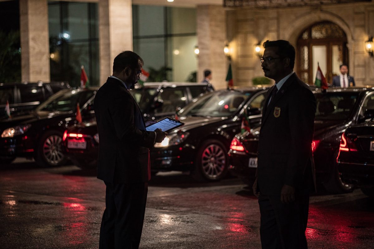 At night on a New Delhi hotel driveway, a pair of security staff in dark suits stand beside a line of black cars with small flags, one holding a clipboard at.