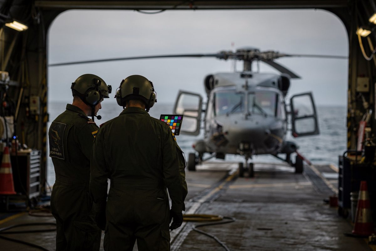 Wide shot inside a helicopter hangar at first light with the doors rolled open onto grey water, two aircrew in flight suits seen from behind studying a small.