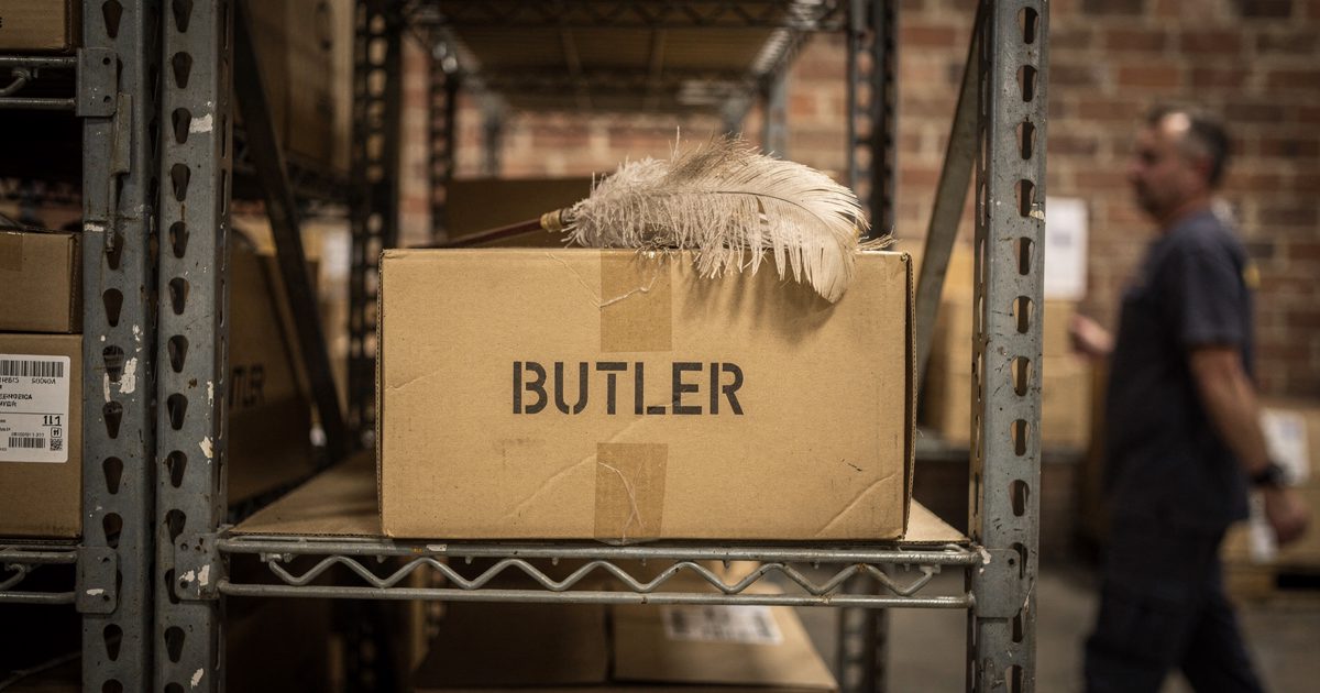 Close detail in flat midday light between tall steel pallet racks in a suburban warehouse, a cardboard archive box stencilled "BUTLER" on a shelf with a.