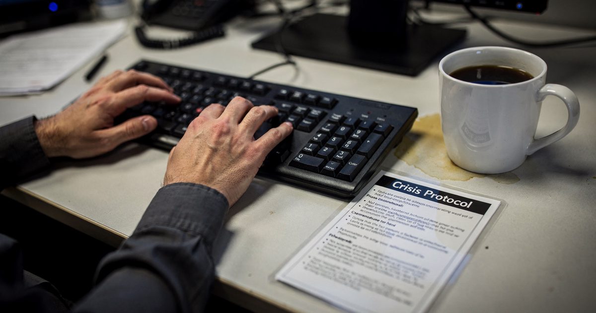 Close-up of hands typing on a computer keyboard under harsh fluorescent office lights, with a laminated 'Crisis Protocol' card resting next to a half-empty.