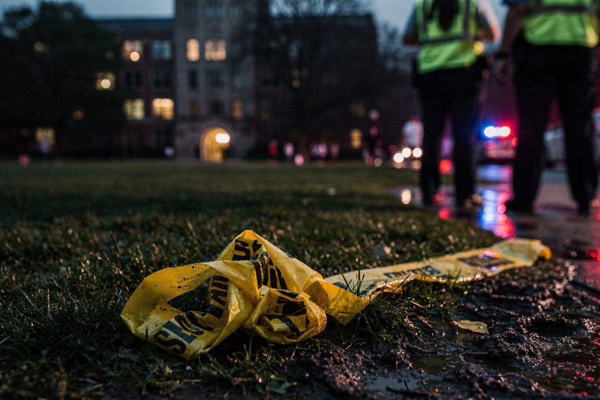 Discarded yellow police tape tangled in the wet grass of a university quadrangle at dusk, illuminated by the red and blue flashing lights of out-of-frame.