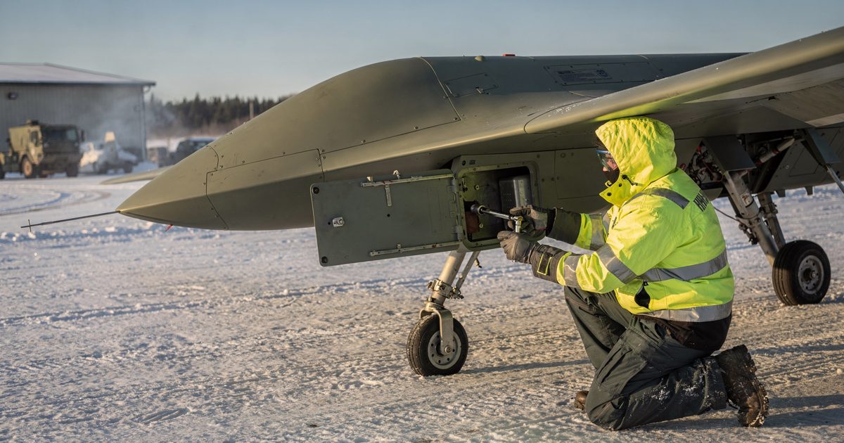 Harsh afternoon sunlight across a snow-covered tarmac at a rural airbase as a ground crew mechanic in a high-visibility parka uses a socket wrench to tighten a.