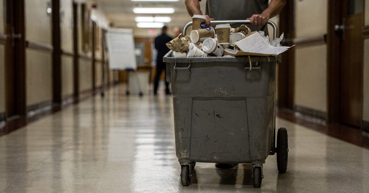 Harsh fluorescent light reflecting off a polished linoleum courthouse corridor at midday, a maintenance worker seen from the shoulders down dragging a heavy.