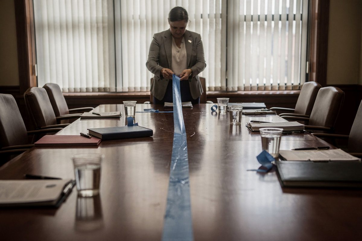 Flat afternoon daylight filtering through vertical blinds in a deserted deliberation room, a court clerk framed from the waist down peeling a thick dividing.
