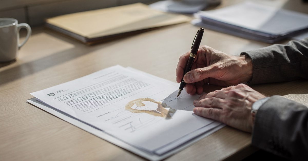 Close crop of a civil servant's hands at a Geneva desk in hard late-morning window light, filling a printed two-page form with a fountain pen, a coffee ring.