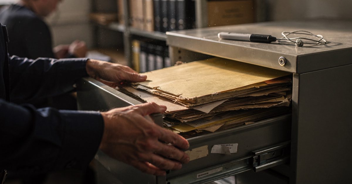 Close-up of an archivist's hands sliding a bulging manila folder into an overstuffed metal filing drawer in a records room, late afternoon light slanting.