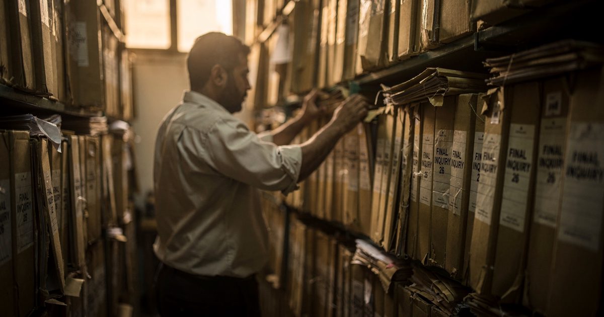 Warm late-afternoon light in a narrow Cairo ministry records aisle as a clerk cropped at the chest, face turned to the shelves, slides one more brown folder.