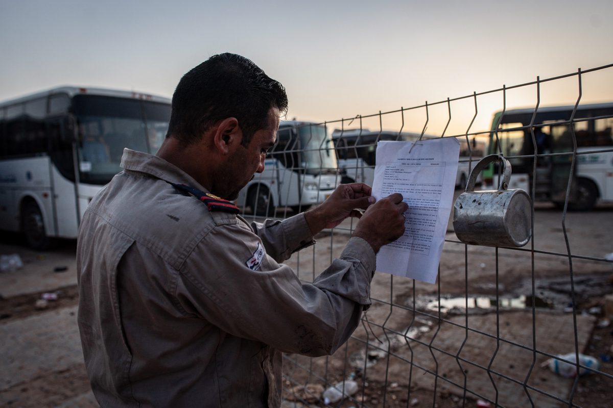 Pale first light in a south Sinai coach depot yard as a driver cropped at the chest, head bent, pins a typed sheet to a wire fence beside idle coaches, a tin.