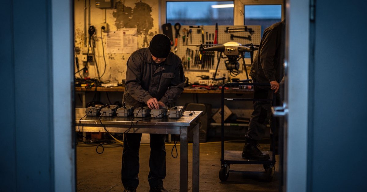 Through a doorway in a Tallinn drone workshop under amber evening lamps, a figure in a wool cap bends to line six drone batteries along a steel bench while a.