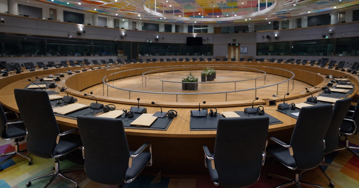 Wide news photograph of an anonymous European government conference room with empty chairs, beige folders, translation headsets and a circular meeting table.