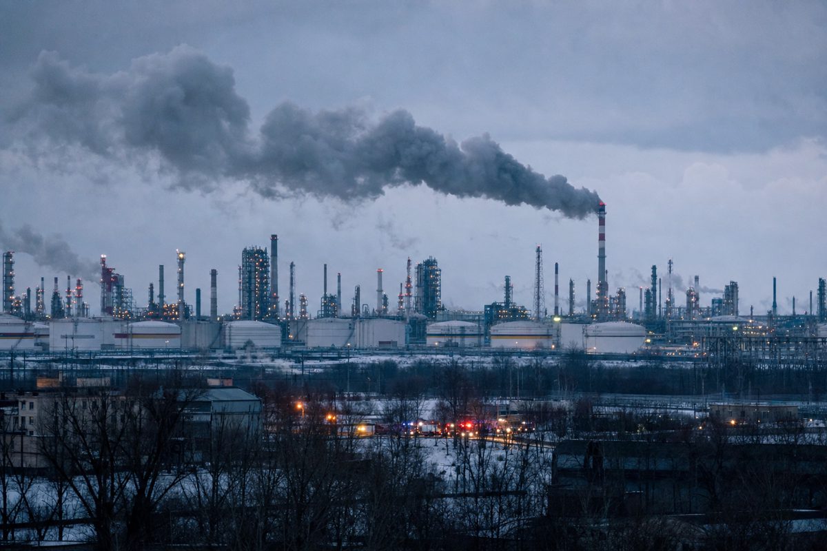 Distant documentary photograph of an industrial oil refinery skyline at dusk, storage tanks and pipework beneath a smoky winter sky, emergency vehicles visible.