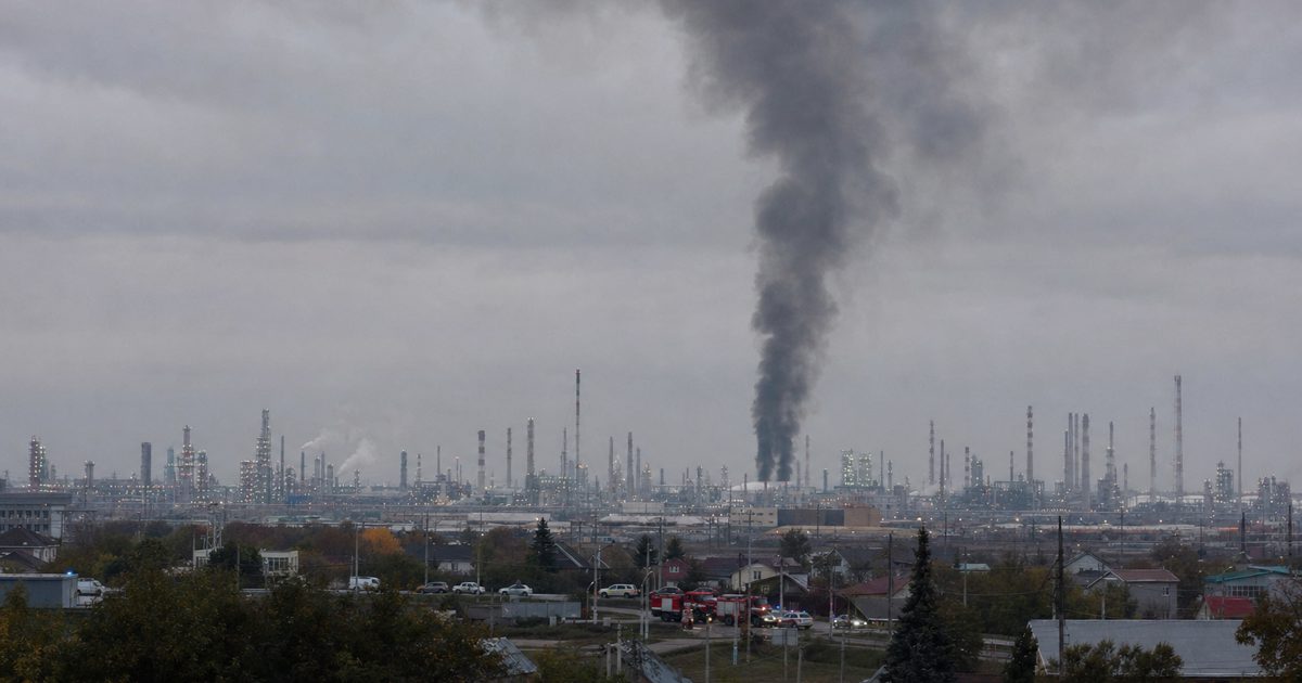 Wide photojournalistic view of a distant industrial refinery skyline beneath a grey sky, a column of smoke rising in the background, emergency vehicles small.