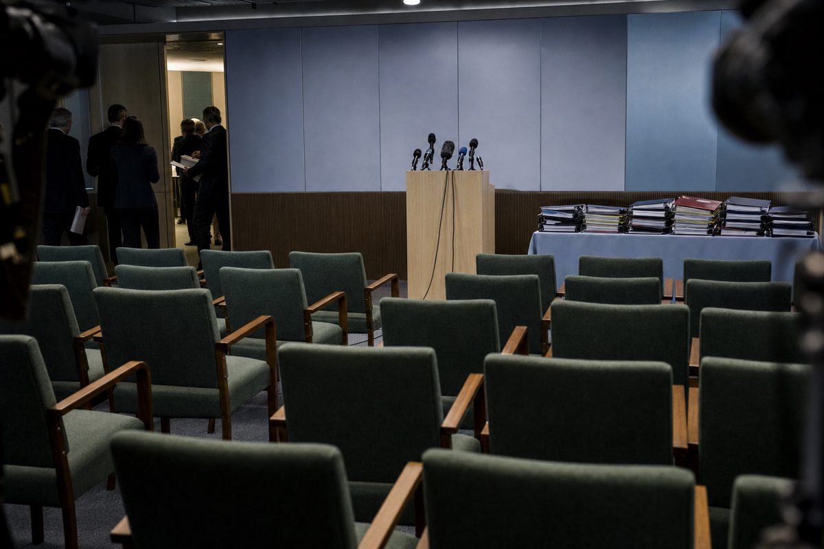 Quiet international press briefing room with rows of empty chairs, a plain lectern, folders and microphones under cool overhead lighting, anonymous staff.
