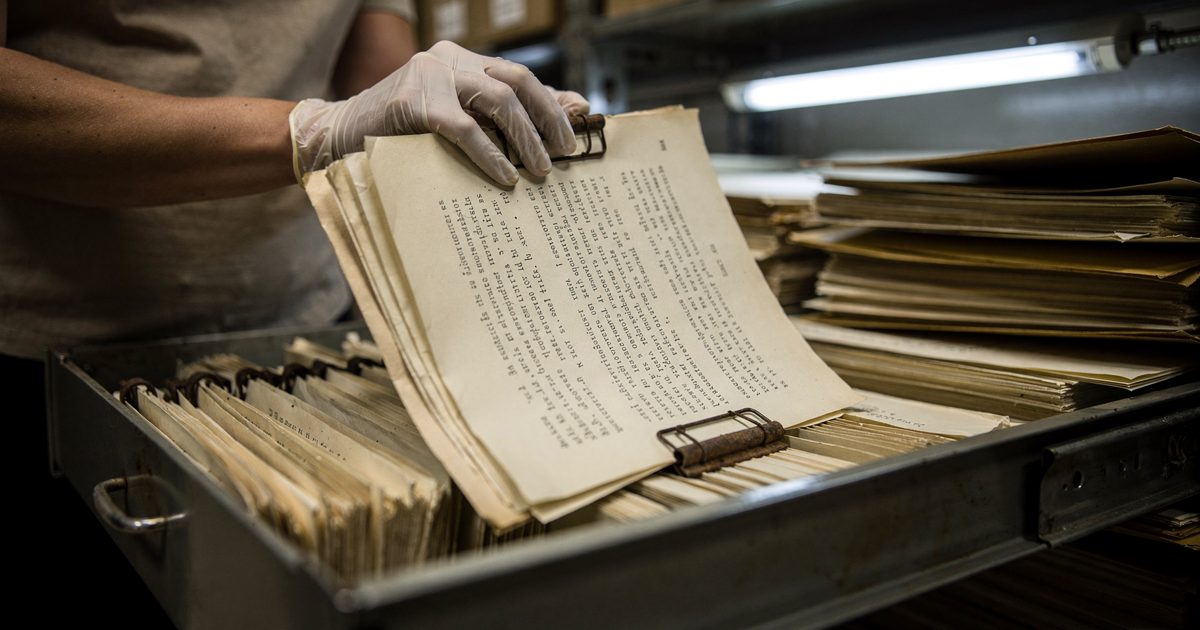 Close-up of gloved hands lifting a thick typed treaty draft from a metal filing drawer in a strip-lit basement archive, the pages held together by a rusting.
