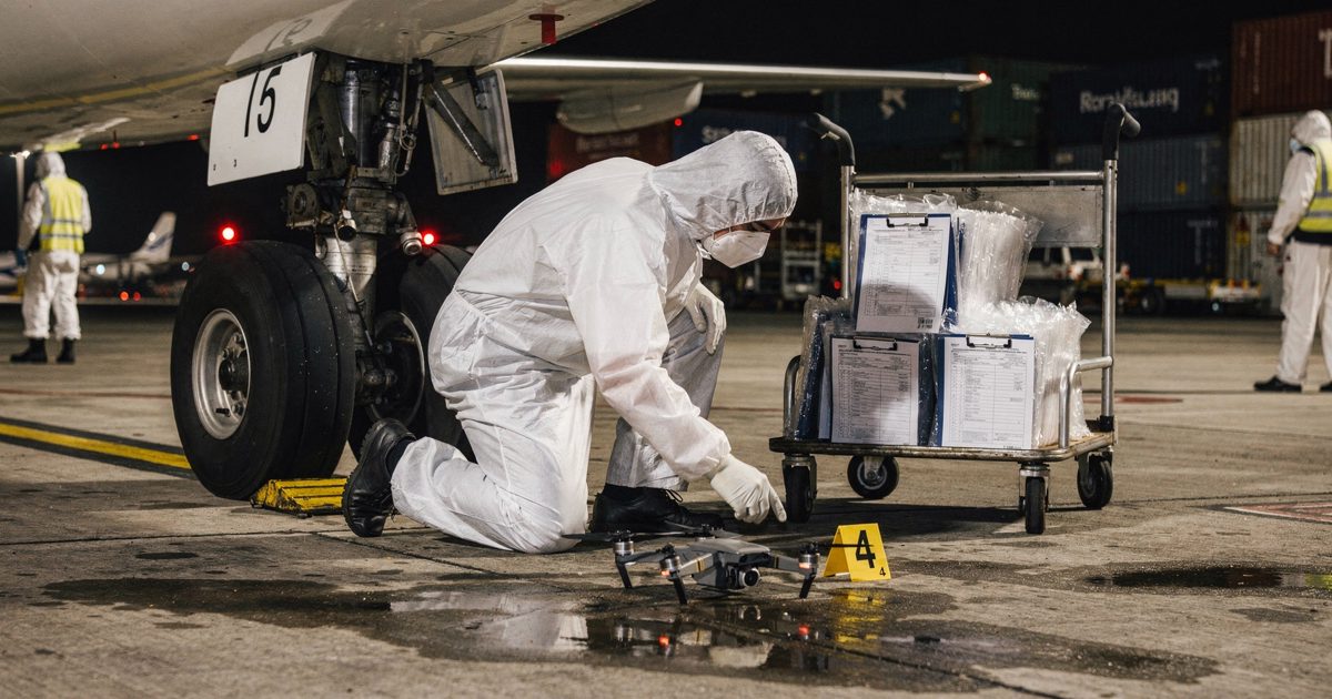Floodlit night on a cargo airport apron, a forensic investigator in a white coverall kneeling at the nose wheel of a freight jet, placing a numbered yellow.
