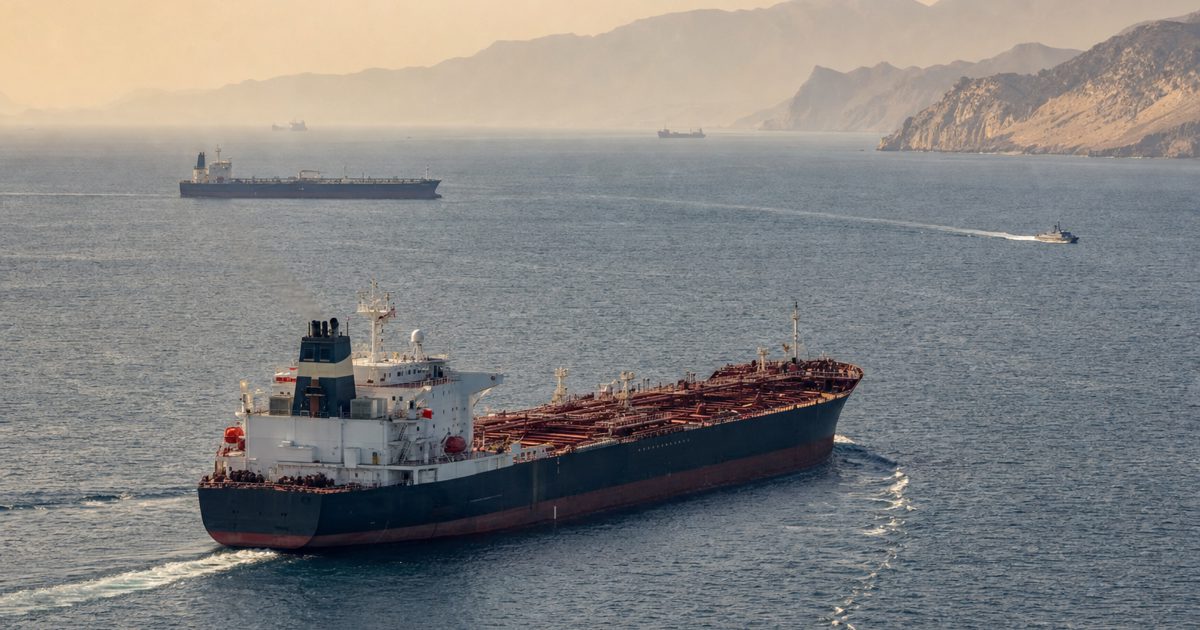 Wide documentary news photograph of cargo tankers moving through the Strait of Hormuz at hazy dawn, with rugged coastline in the distance and a small patrol.