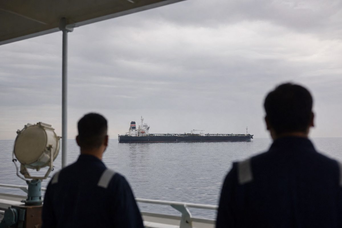 Long-lens photograph from a commercial ship’s deck showing another tanker crossing calm Gulf waters under an overcast sky, anonymous crew silhouettes viewed.