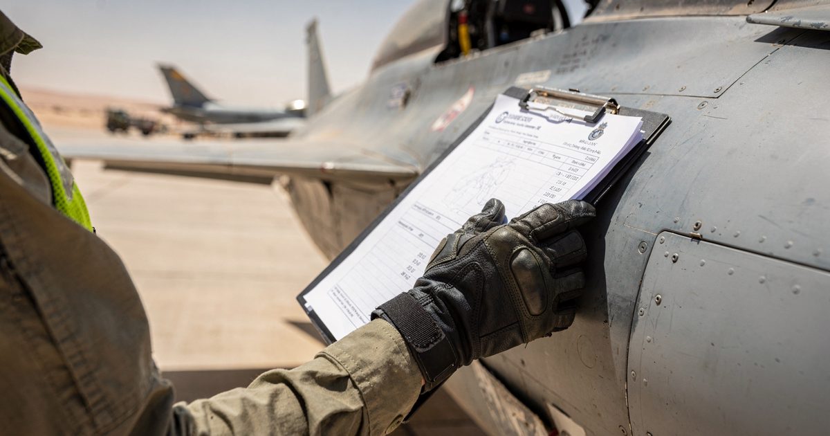 Tight close-up at blazing noon on a desert airbase apron, a gloved RAF ground-crew hand pressing a clipboard flat against the grey fuselage of a Typhoon.