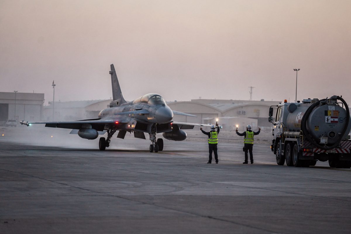 Wide shot at pale dawn across a Gulf air base runway, a single RAF Typhoon taxiing through rising heat haze while two small silhouetted ground crew wave it.