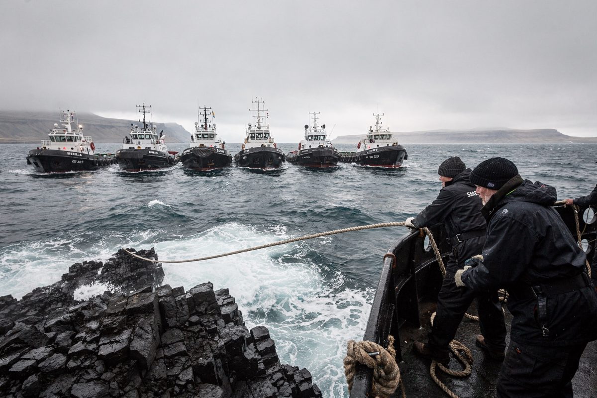 Pale midday glare on a black lava shoreline where a line of tugs holds station just beyond the surf and crew on the nearest deck, faces unreadable under wool.