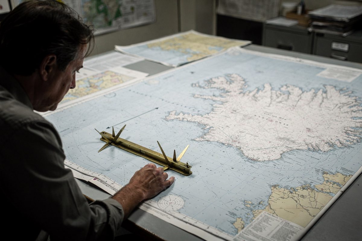 Cool overhead office light in a government map room as a civil servant seen in side profile with face tipped to the table sets a brass chart divider on a.