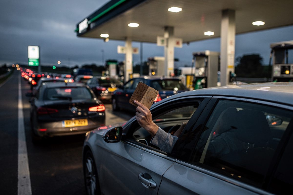 Wide shot at dusk across a motorway services forecourt with every pump lit, cars queued nose to tail down the slip road and one driver's arm hanging from a.