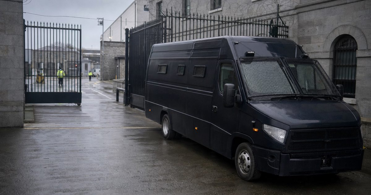 A documentary news photograph of an unmarked dark armoured transport van parked inside a secured government vehicle compound in Dublin, guarded gates and wet.