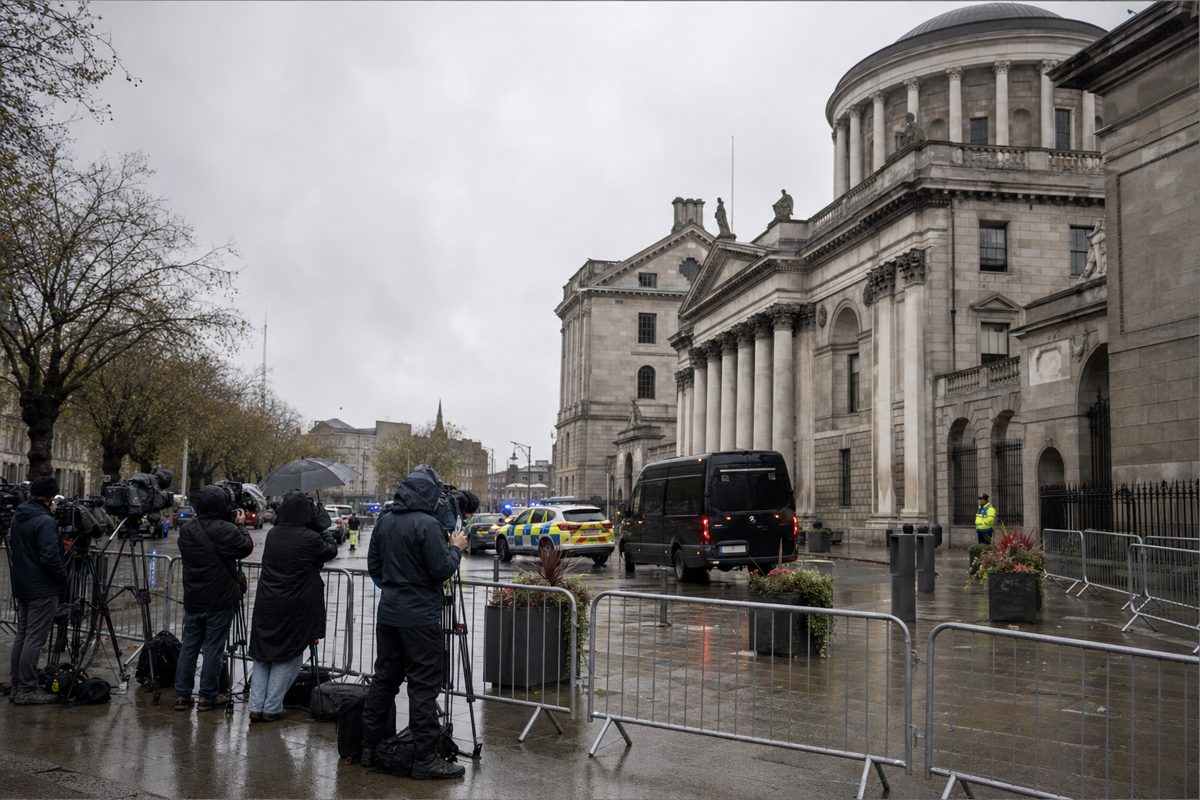 A wide news photograph of a courthouse entrance on a rainy morning with temporary metal barriers, police vehicles in the distance and journalists waiting.